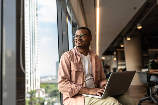 A man sitting by a window with a laptop on his lap, looking outside and smiling